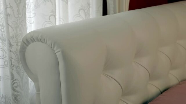 Beautiful white leather headboard upholstered in classic style (capitone) in bedroom with burgundy walls next to window with tulle and curtains. Shot in motion