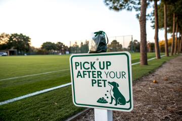 A sign instructing pet owners to clean up after their dogs is placed beside a grassy field, promoting responsible pet ownership in a park setting.