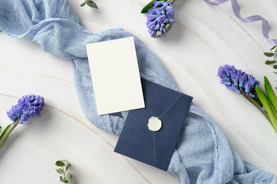 Luxury wedding stationery set with flowers. Elegant flat lay of a blank white card and navy blue envelope with a wax seal, surrounded by purple hyacinth flowers and a light blue fabric on marble table