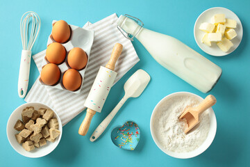 Baking ingredients and tools on a blue background, including eggs, flour, butter, milk, a whisk, a rolling pin, and cookie cutter. Flat lay, top view