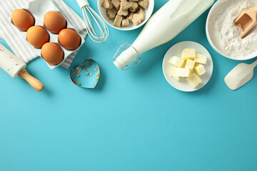 Baking ingredients on a blue background: eggs, milk, butter, flour, sugar cubes, whisk, rolling pin, and sprinkles. Flat lay, top view