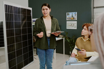 Asian woman engaging audience with solar panel details in modern office setting holding tablet...