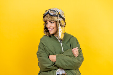 Young beautiful aviator woman wearing vintage pilot helmet with glasses and jacket over yellow background looking to the side with arms crossed convinced and confident