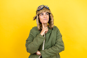 Young beautiful aviator woman wearing vintage pilot helmet with glasses and jacket over yellow background serious face thinking about question with hand on chin, thoughtful about confusing idea