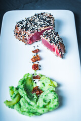 Tuna slices with green puree, sesame seeds and sun-dried tomatoes on a white plate on a dark...