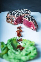 Tuna slices with green puree, sesame seeds and sun-dried tomatoes on a white plate on a dark...