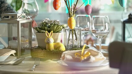 A young woman serves a festive Easter table with a yellow bunny decoration. Sunlight fills the cozy room, highlighting elegant tableware, pastel colors, and holiday decor. A warm family moment full of