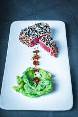 Tuna slices with green puree, sesame seeds and sun-dried tomatoes on a white plate on a dark...