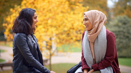 Two Young British Muslim Female Friends Sitting On Bench In Autumn Park