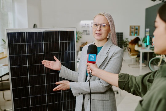 Woman in professional attire discussing solar panel technology during TV interview while holding microphone. Presenter in background holding another microphone to elicit responses