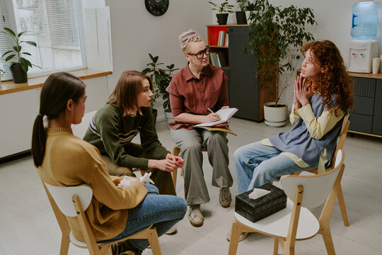 Young adults sitting in circle, actively participating in group therapy session, supporting each other, while counselor listens attentively and takes notes