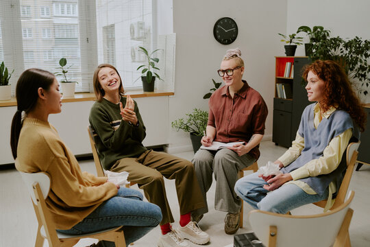 Group of women seated in circle discussing personal topics in cozy therapy room surrounded by plants and light from large windows. Sharing experiences and support in comfortable environment - Powered by Adobe