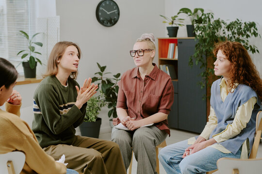 Group therapy session with four individuals discussing mental health issues. Participants sharing personal experiences in supportive environment with plants and clock in background