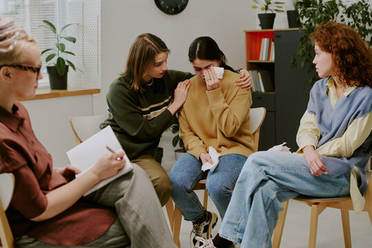 Counselor holding group therapy for emotional support while one participant comforting another with gentle hug holding tissue. Discussing experiences and feelings in warm setting