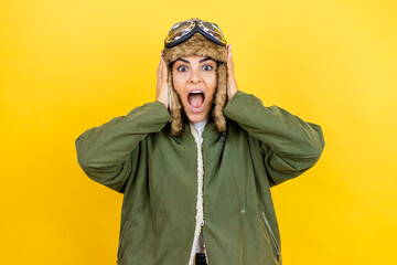 Young beautiful aviator woman wearing vintage pilot helmet with glasses and jacket over yellow background crazy and scared with hands on head, afraid and surprised of shock