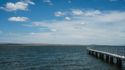 Fototapeta premium A long wooden dock extends into a calm lake on a sunny day. The water is a deep blue-green color and there are ripples on the surface. The sky is clear with a few white clouds.