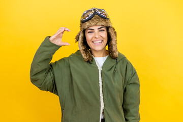 Young aviator woman wearing vintage pilot helmet with glasses and jacket over yellow background smiling and confident gesturing with hand doing small size sign with fingers . Measure concept.