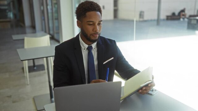 Young man working indoors in an office, holding a notepad and pen, dressed in a suit, focused on his laptop on a modern desk with indoor lighting