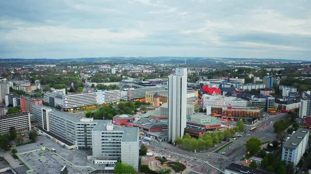 Aerial footage of Chemnitz Stadthalle surrounded by tall buildings under a cloudy sky, showcasing urban architecture and city dynamics. Drone move forward