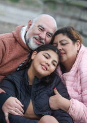 Obraz premium portrait of happy latino family, mature couple with teenage girl, hugging outdoors sitting on stairs on cloudy cold day