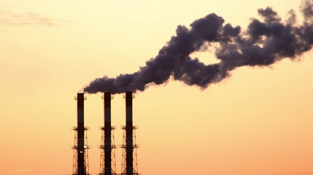 Factory chimneys emit thick black smoke into the sky against the backdrop of a sunset. Environmental issue, air pollution, industrial landscape.
