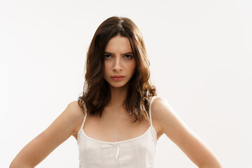 Obraz premium Young brunette woman in a white light top posing against a white background, expressing emotions and touching her face with her hands. Minimalistic and natural beauty concept
