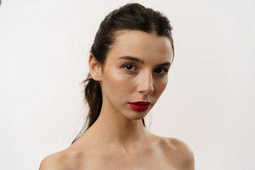 Obraz premium Young brunette woman with red lipstick posing against a white background, expressing emotions, and touching her face with her hands. Minimalistic beauty and fashion concept