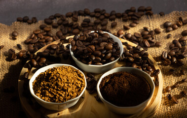 Top view of coffee beans scattered next to small porcelain bowls with different types of coffee. beans, ground and instant, on a wooden plate on a dark background with sunlight coming