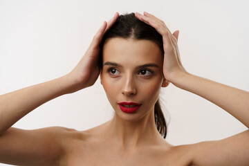 Young brunette woman with red lipstick posing against a white background, expressing emotions, and touching her face with her hands. Minimalistic beauty and fashion concept