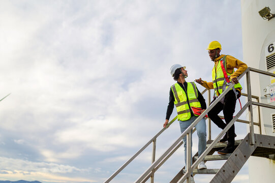 Young black worker and indian engineer wearing safety vests and hard hats discussing maintenance while climbing stairs on a wind turbine