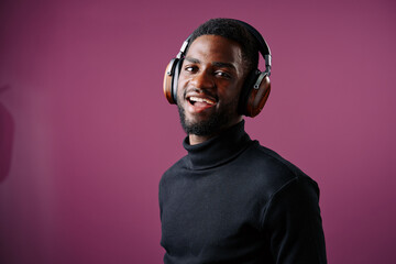 Smiling young Black man wearing headphones against a purple background, showcasing joy and engagement with music in a modern, stylish look