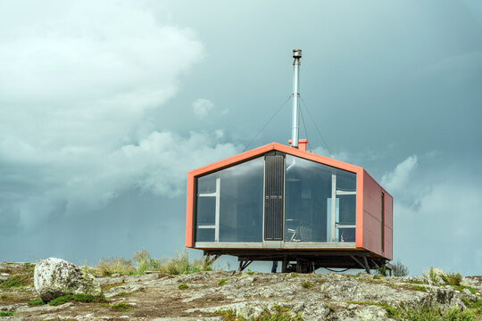 Modern red cabin stands on rocky terrain under dramatic cloudy sky in a remote location