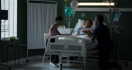 Obraz premium African child in a hospital ward is visited by her dad and mom with balloons and a cake to wish her a happy birthday.