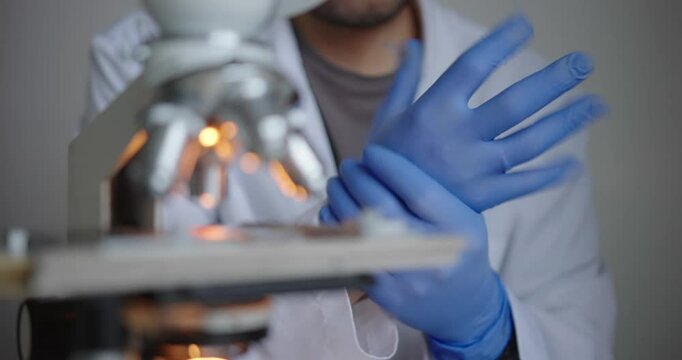 Scientist, dressed in white lab coat, carefully puts on blue gloves as they prepare to examine a microscope