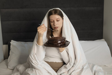 Upset sad worried woman eating sweet cake. Wrapped in blanket in the bedroom, eating personal...