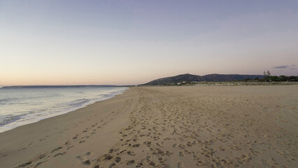 A solitary figure walks along a deserted beach at sunrise. The sand is soft and golden, and the waves gently lap at the shore. In the distance, a line of trees marks the horizon.