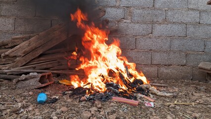 Vibrant flames consume a pile of refuse in an open-air setting, set against a rough brick wall. A...