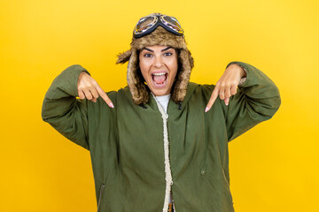 Fototapeta premium Young beautiful aviator woman wearing vintage pilot helmet with glasses and jacket over yellow background looking confident with smile on face, pointing oneself with fingers proud and happy.