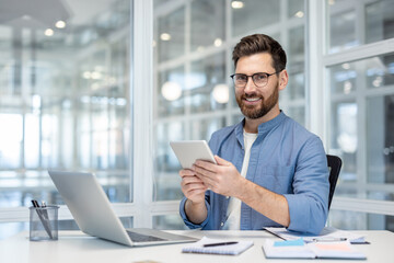 Fototapeta premium Portrait of a successful young businessman, a man is smiling and looking at the camera with a tablet computer in his hands, he is using a laptop at his workplace, working inside the office.