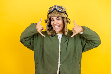 Fototapeta premium Young beautiful aviator woman wearing vintage pilot helmet with glasses and jacket over yellow background shouting with crazy expression doing rock symbol with hands up