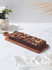 A rich chocolate brownie adorned with almonds is placed on a wooden serving board next to a bouquet of flowers and a silk sheet