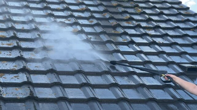 A person using a high-pressure washer to clean a dirty tiled roof, removing moss and dirt buildup with a strong water spray