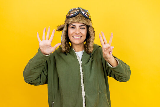 Young beautiful aviator woman wearing vintage pilot helmet with glasses and jacket over yellow background showing and pointing up with fingers number eight while smiling confident and happy