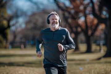 Fototapeta premium Smiling man jogging in a park on a sunny day, wearing a dark athletic top and headphones, with trees and grass in the background.