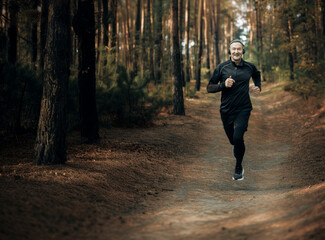 Fototapeta premium Man jogging on a peaceful forest path, wearing black athletic gear and headphones, surrounded by tall trees and a tranquil natural atmosphere