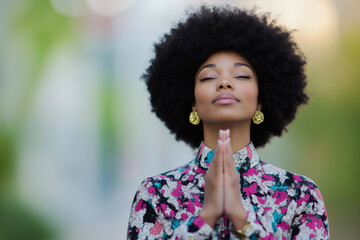 A black woman with afro hair and elegant gold earrings meditating outdoors, wearing a colorful floral blouse with her hands in a prayer position and eyes closed