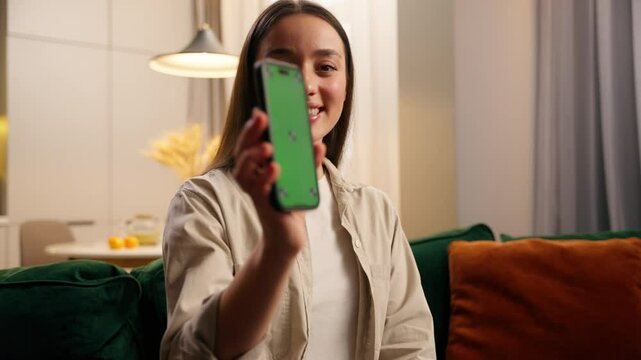 Caucasian woman female lady girl at home relaxing at couch holding mobile phone smiling browsing social media app showing green screen smartphone display mock up chroma key cellphone website internet
