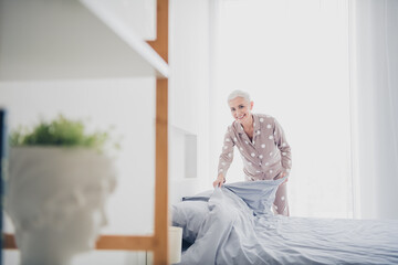 Charming elderly woman in pajamas making bed in a bright room, enjoying a peaceful morning with natural light creating a cozy atmosphere