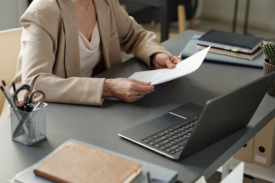 Hands of mature businesswoman with new contract looking through its terms while sitting by workplace