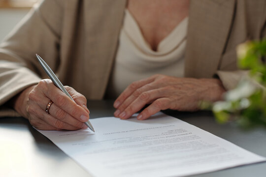 Hand of aged female agent or lawyer with silver pen over contract during signing financial document by desk